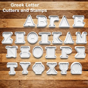 May include: White plastic Greek letter cutters and stamps arranged on a wooden surface. The text "Greek Letter Cutters and Stamps" is at the top. The letters are in a sans-serif font.