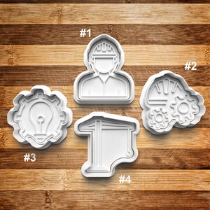 May include: Four white plastic cookie cutters shaped like a construction worker, a gear with a lightbulb, a graduation cap, and a wrench and gears. The cutters are numbered 1 through 4.