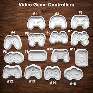 May include: A collection of fifteen white plastic cookie cutters shaped like various video game controllers and handheld consoles, labeled with numbers one through fifteen, arranged on a dark brown wood grain surface under the text Video Game Controllers.