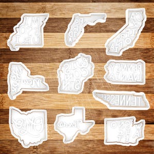 May include: White cookie cutters shaped like the outlines of different US states. Each state has the state name spelled out in white letters inside the outline. The cookie cutters are on a wooden surface.