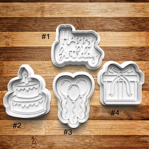 May include: Four white plastic cookie cutters on a wooden surface. The cutters are shaped like a birthday cake, balloons, a gift box, and the words "Happy Birthday". Each cutter is labeled with a number.