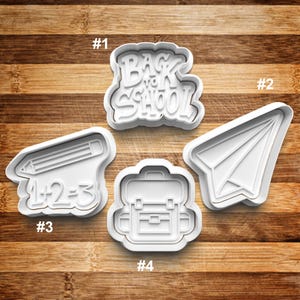 May include: Four white plastic cookie cutters shaped like a pencil with the equation 1+2=3, a backpack, a paper airplane, and the words "Back to School".