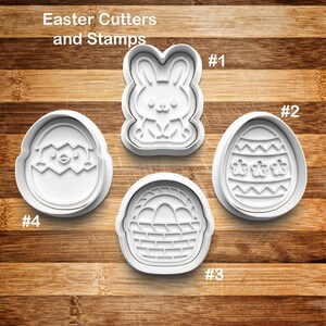 May include: Four white plastic Easter cookie cutters and stamps on a wooden surface. The cutters include a bunny, an egg with floral designs, a basket with eggs, and a chick hatching from an egg. The text "Easter Cutters and Stamps" is at the top.