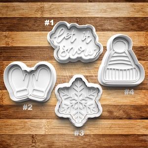May include: Four white plastic cookie cutters on a wooden surface. The cutters are shaped like mittens, a snowflake, a hat, and a banner that says "Let it snow".