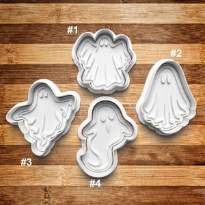 May include: Four white plastic ghost-shaped cookie cutters. Each cutter has a different ghost design, including a ghost with wings, a classic ghost shape, and two other unique ghost forms. The cutters are on a wooden surface.
