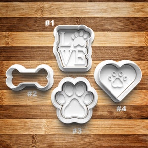 May include: Four white plastic cookie cutters on a wooden surface. The cutters are shaped like a bone, a paw print, a heart with a paw print inside, and a rectangle with the word "LOVE" and a paw print inside. The cutters are numbered 1 through 4.