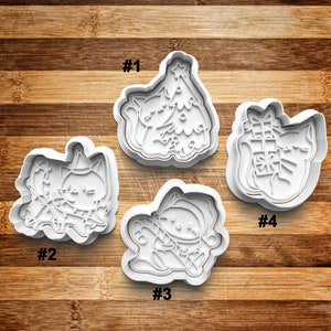 May include: Four white plastic cookie cutters shaped like cats in various Christmas-themed poses. The cutters are numbered 1 through 4. Cutter #1 shows a cat holding a Christmas tree. Cutter #2 shows a cat wearing a party hat and holding a candy cane. Cutter #3 shows a cat wearing a Santa hat and holding a candy cane. Cutter #4 shows a cat holding a gift box.