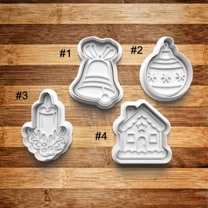 May include: Four white plastic Christmas cookie cutters. The cutters are shaped like a bell, an ornament, a candle, and a gingerbread house. The cutters are numbered 1 through 4.