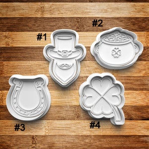 May include: Four white plastic cookie cutters shaped like a leprechaun, a pot of gold, a horseshoe, and a four-leaf clover. The cutters are numbered 1 through 4.