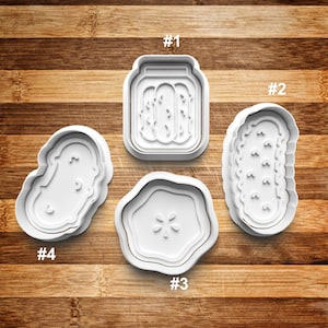 May include: Four white plastic cookie cutters shaped like a jar of pickles, a pickle, a pickle slice, and a pickle. The cutters are numbered 1 through 4.