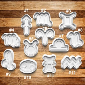 May include: A collection of twelve white plastic cookie cutters, each representing a different zodiac sign. The cutters are arranged on a wooden surface and labeled with numbers from #1 to #12.