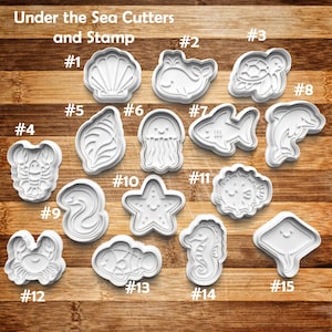 May include: A collection of white plastic cookie cutters in various sea creature shapes, including a seashell, whale, turtle, lobster, jellyfish, fish, dolphin, eel, starfish, and seahorse. The text "Under the Sea Cutters and Stamp" is at the top.