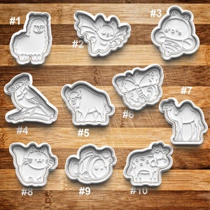May include: Ten white plastic cookie cutters shaped like various animals and insects, including an alpaca, bat, bee, blue jay, buffalo, butterfly, camel, cat, fish, and cow. The cutters are numbered 1 through 10.