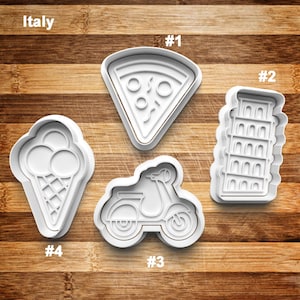 May include: White plastic cookie cutters in the shapes of a pizza slice, ice cream cone, scooter, and the Leaning Tower of Pisa. The word "Italy" is above the cutters. The cutters are on a wooden surface.