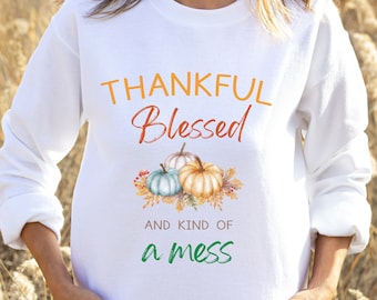 May include: White sweatshirt with the words "THANKFUL Blessed AND KIND OF a mess" in orange, red, and green. The design includes three pumpkins in shades of blue, white, and orange with fall foliage.