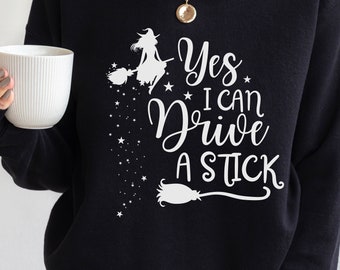 May include: Black sweatshirt with the text "Yes I Can Drive A Stick" in white script. A white witch on a broom and stars are also featured in the design. A fun Halloween-themed top.