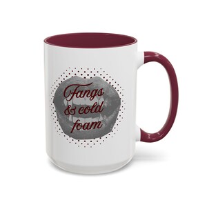 May include: White ceramic mug with a claret interior and handle. The mug features a graphic of a mouth with fangs and the text "Fangs & cold foam" in claret. The design is surrounded by a dotted circle.