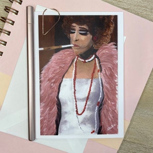 May include: An art print of a person with dark hair and makeup, wearing a white dress and a pink feather boa, smoking a cigarette. The print is on a white card with the artist's signature. A rose gold pen and a heart-shaped paper clip are next to the card.