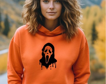 May include: Orange hoodie featuring a black graphic of a Scream mask. The hoodie has a front pocket and a drawstring hood. The design is a spooky, Halloween-themed graphic. The hoodie is made of a soft material.