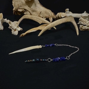 May include: A white bone pendant with a silver chain and blue and black beads. The pendant is shaped like a long, thin blade.