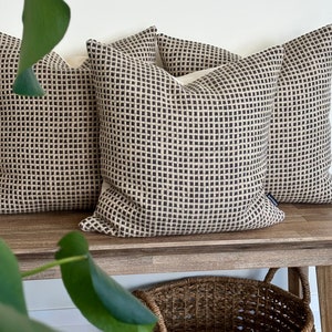 May include: Three brown and beige patterned throw pillows with a subtle grid design. The pillows are arranged on a wooden bench.