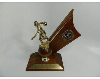 May include: A gold-colored trophy with a wooden base. The trophy features a female figure bowling a ball. The base has a plaque that reads "Northwest Ladies Classic 68 Champion 71".