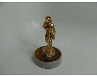May include: A gold-colored figurine of a person in a flowing dress, mounted on a round base with a gray border. The figurine is detailed and appears to be made of metal. The base has text around the edge.