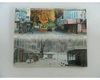 May include: A calendar featuring a collage of four images of a small town in different seasons. The top image shows a street scene with trees in autumn colors. The bottom image shows the same street scene covered in snow. The text "Seasons of Mackinac - 2016 Calendar" is printed at the bottom of the image.