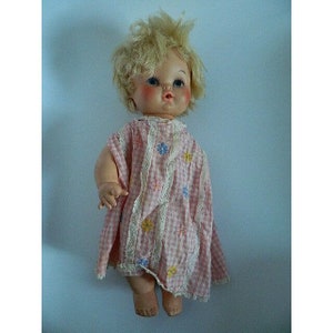 May include: Vintage baby doll with blonde hair and rosy cheeks, wearing a pink and white checkered dress with floral accents and lace trim. The doll has blue eyes and is in a standing position.