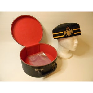 Puede incluir: Una caja de sombrero negra con un interior rojo y un cierre de cremallera. La caja tiene un asa y está abierta para revelar una bolsa de plástico transparente en el interior. Un sombrero negro con ribetes dorados y un diseño de águila bordado está sentado sobre una cabeza de maniquí blanca.