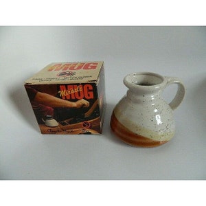 May include: A white ceramic mug with brown speckles and a brown stripe around the middle. The mug is on a white surface. The box in the background says "Miracle Mug" and "Clay to Finish".