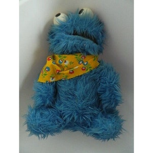 May include: A plush blue monster with large white eyes and a yellow bandana with a floral pattern. The toy has shaggy fur and is a popular children's character.