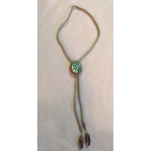 Op de afbeelding: Een zilveren bolo stropdas met een turquoise ovale middenstuk. Het middenstuk is omgeven door een zilveren rand en heeft een textuur, mozaïekachtig uiterlijk. De stropdas heeft een zilveren koord en decoratieve zilveren uiteinden.