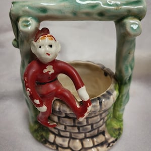 May include: A ceramic figurine of an elf sitting on the edge of a well. The well is made of brown and gray bricks and has a green and white wooden frame. The elf is wearing a red suit with white accents.