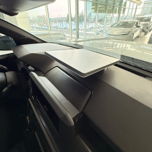 May include: A light gray, rectangular tray is mounted on the dashboard of a vehicle. The tray is positioned above a dark gray, textured dashboard. The interior of the car is visible, with a view of the outside through the windshield.
