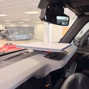 May include: A gray, rectangular tray mounted on a vehicle's dashboard. The tray is positioned above the dashboard controls and is near the rearview mirror. The interior of the vehicle is visible through the windshield.