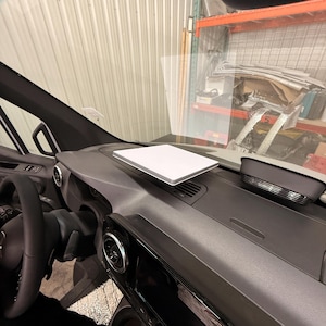May include: A white, rectangular device is placed on the dark grey dashboard of a vehicle. The black steering wheel and interior details are visible. The background shows a glimpse of a warehouse setting.