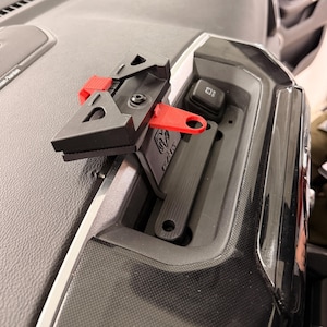 May include: Black and red car dashboard phone mount. The mount is designed to fit into a vehicle's dashboard. The mount has a red lever and the RAM logo is visible on the mount.