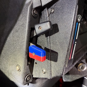 Könnte beinhalten: Nahaufnahme eines schwarzen Kunststoffpanels mit zwei blau-roten Kippschaltern und einem schwarzen Kippschalter. Das Panel hat eine silberne Schraube und ein Schlüsselloch.