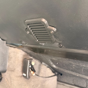 May include: A black plastic car vent cover with horizontal slats, mounted on a dark surface with two screws. The vent is rectangular with rounded edges. The image shows a close-up of the vent and surrounding area.