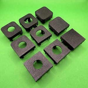 May include: Nine black, square plastic components with a textured surface are displayed. Some have a central circular hole, others are solid. The components are arranged on a bright green background.