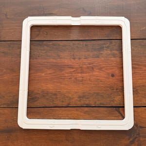 Puede incluir: Un bastidor de bordado cuadrado, de color blanquecino, con un diseño rectangular con esquinas redondeadas. El bastidor está hecho de un material rígido y tiene una serie de pequeñas hendiduras circulares a lo largo de sus bordes. Está diseñado para sujetar la tela tensa para bordar.