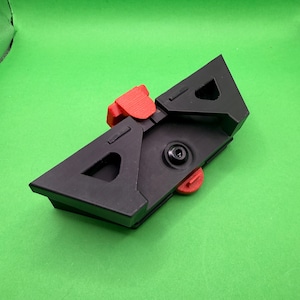 May include: A black plastic mechanical component featuring a triangular frame with two large cutouts. It includes a bright red textured lever at the top and a smaller red circular dial on the underside, secured by a central black metal screw.