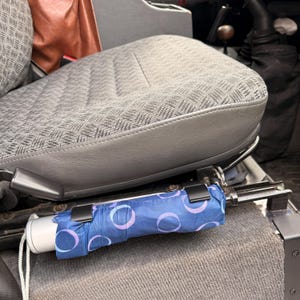 May include: A small, folded umbrella with blue fabric and a pattern of light purple circles. It's attached to a vehicle seat frame with black clips. The seat is grey with a patterned design.