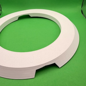 Puede incluir: Objeto circular blanco con superficie texturizada y borde biselado. Presenta dos recortes rectangulares. El objeto está sobre un fondo verde. El diámetro es de aproximadamente 20 cm.