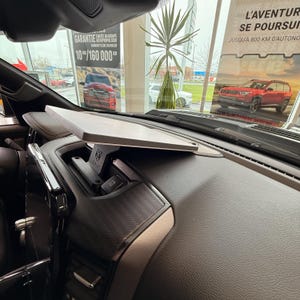 May include: A grey, rectangular tray is mounted on a black bracket, positioned above the centre console of a vehicle. The bracket features the RAM logo. The dashboard is dark brown with a textured pattern. The background includes dealership signage and a red vehicle.