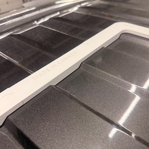 Op de afbeelding: Een donkergrijze, rechthoekige kunststof component met een verhoogde, witte, L-vormige rand. Het witte randgedeelte is voorzien van de in reliëf aangebrachte tekst MADE IN CANADA. Het oppervlak heeft een reeks parallelle, verzonken groeven.