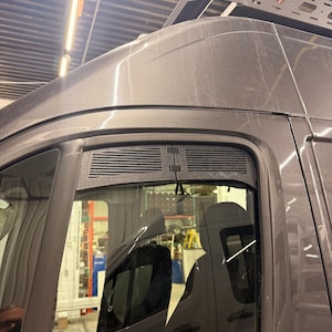 Peut inclure: Un insert de ventilation pour fenêtre de fourgon gris foncé avec une finition en plastique noir texturé et des lamelles horizontales. L'aération rectangulaire est installée le long du bord supérieur du cadre de fenêtre de la porte passager.