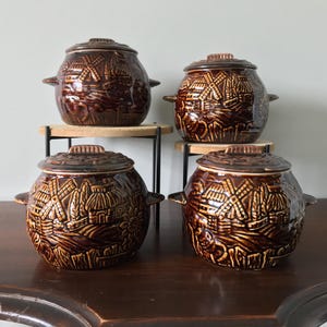 May include: Four brown ceramic lidded pots with handles. Each pot features a detailed relief design of a village scene with windmills. The pots are arranged on a wooden table and small stands.