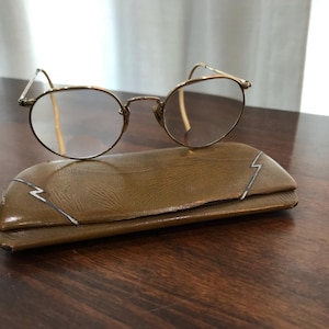 May include: Vintage round eyeglasses with a gold-colored metal frame rest on a brown leather case. The case has a decorative silver lightning bolt design on the front. The glasses have clear lenses.